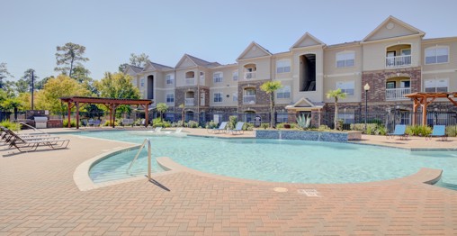 Resort-style outdoor swimming pool surrounded by red brick patio and apartment buildings behind it on a sunny day 